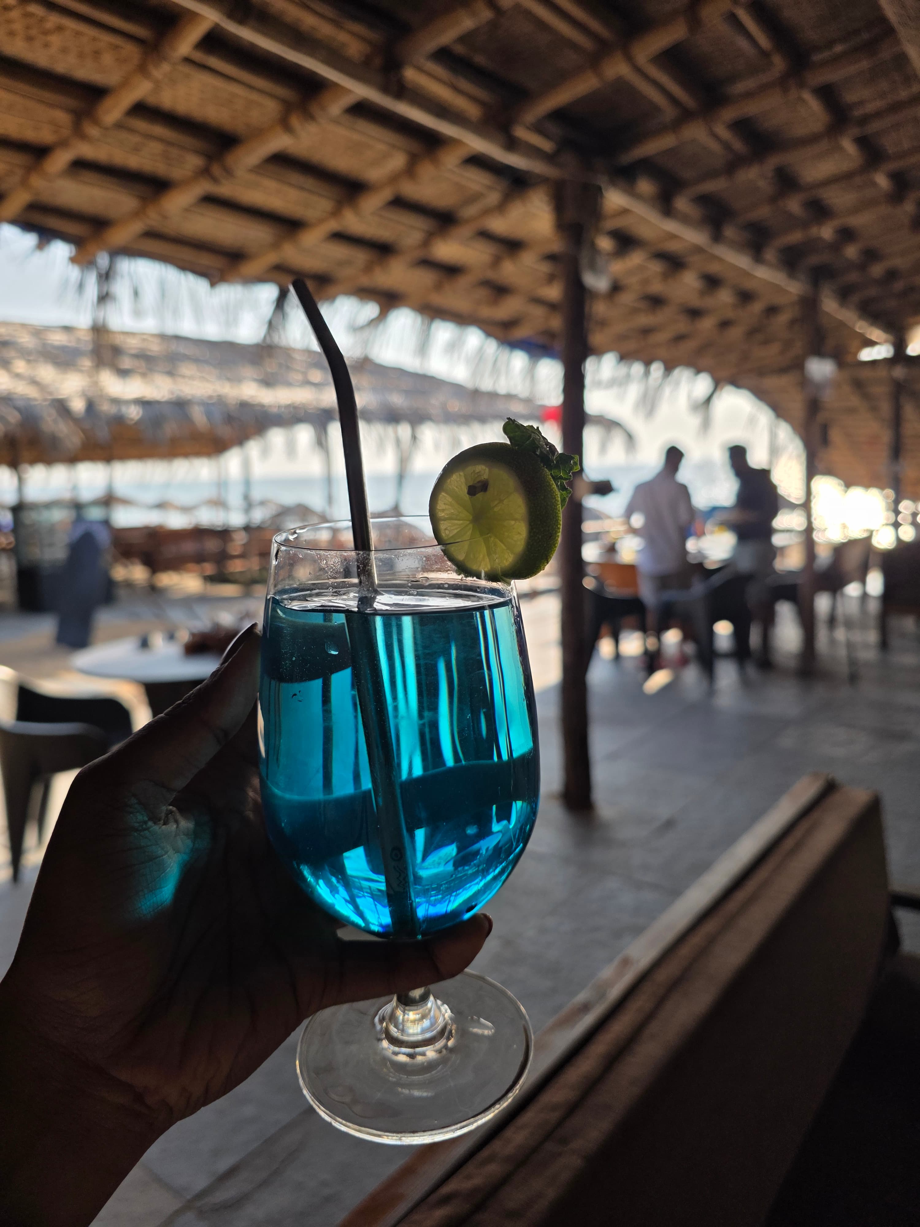 Handcrafted blue lagoon cocktail with lime wheel and black straw held up inside the thatched open-air lounge
