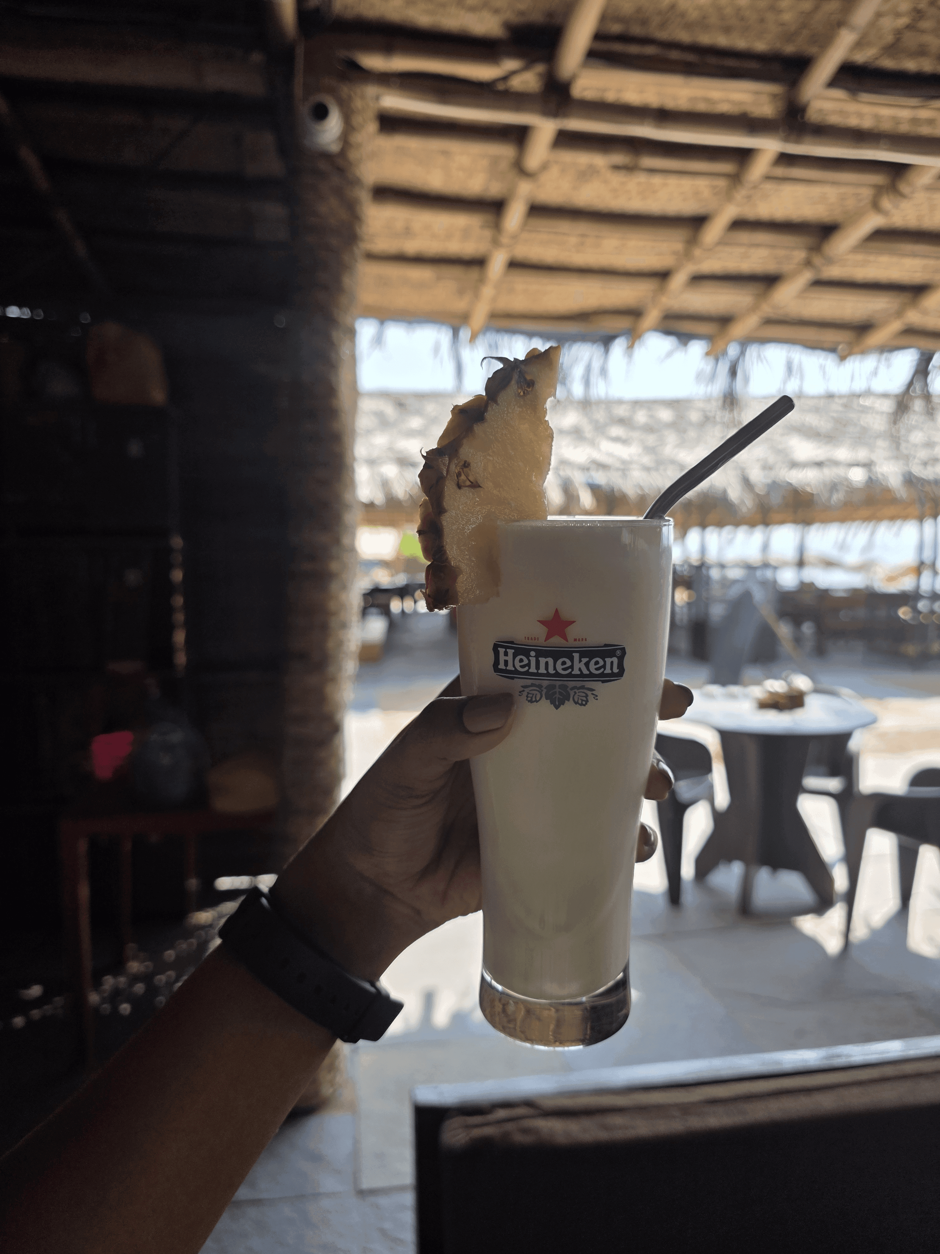 Creamy piña colada with fresh pineapple garnish in a tall Heineken glass held under thatched bamboo ceiling