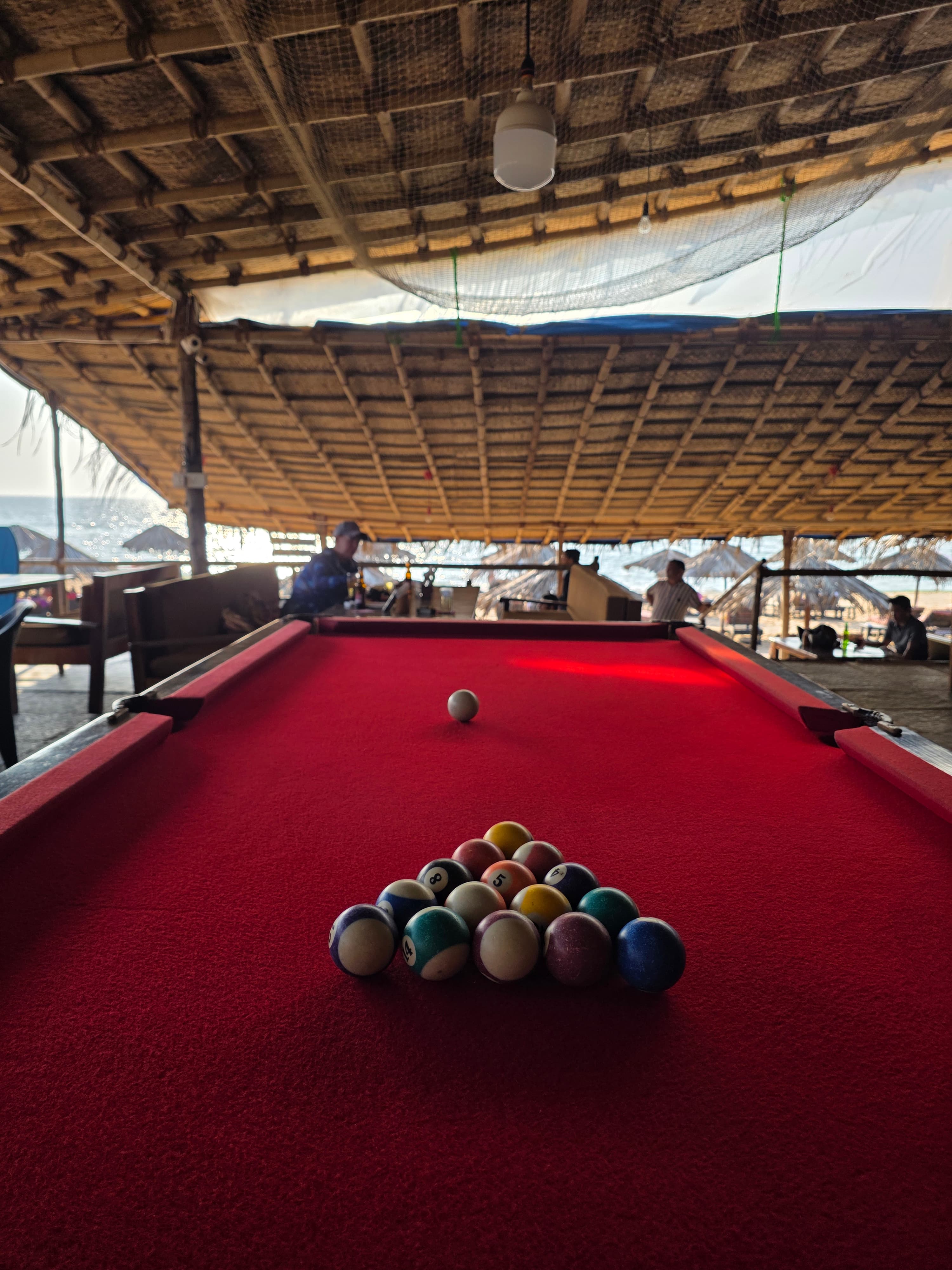 Red felt billiards table set with racked balls, ocean and thatched beach umbrellas visible in the open-air background