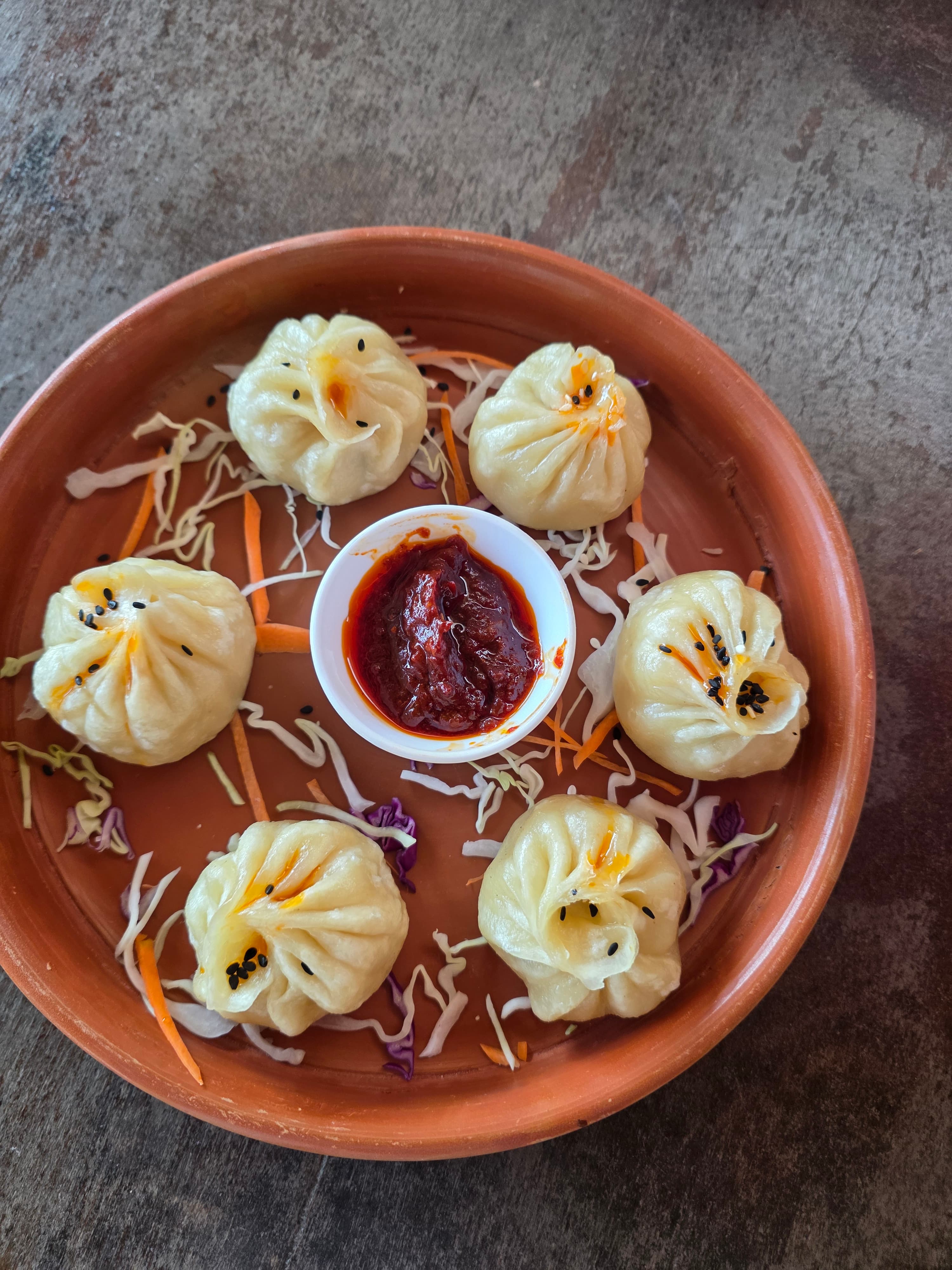 Six steamed momos arranged on a terracotta plate with spicy red chutney dipping sauce in the centre