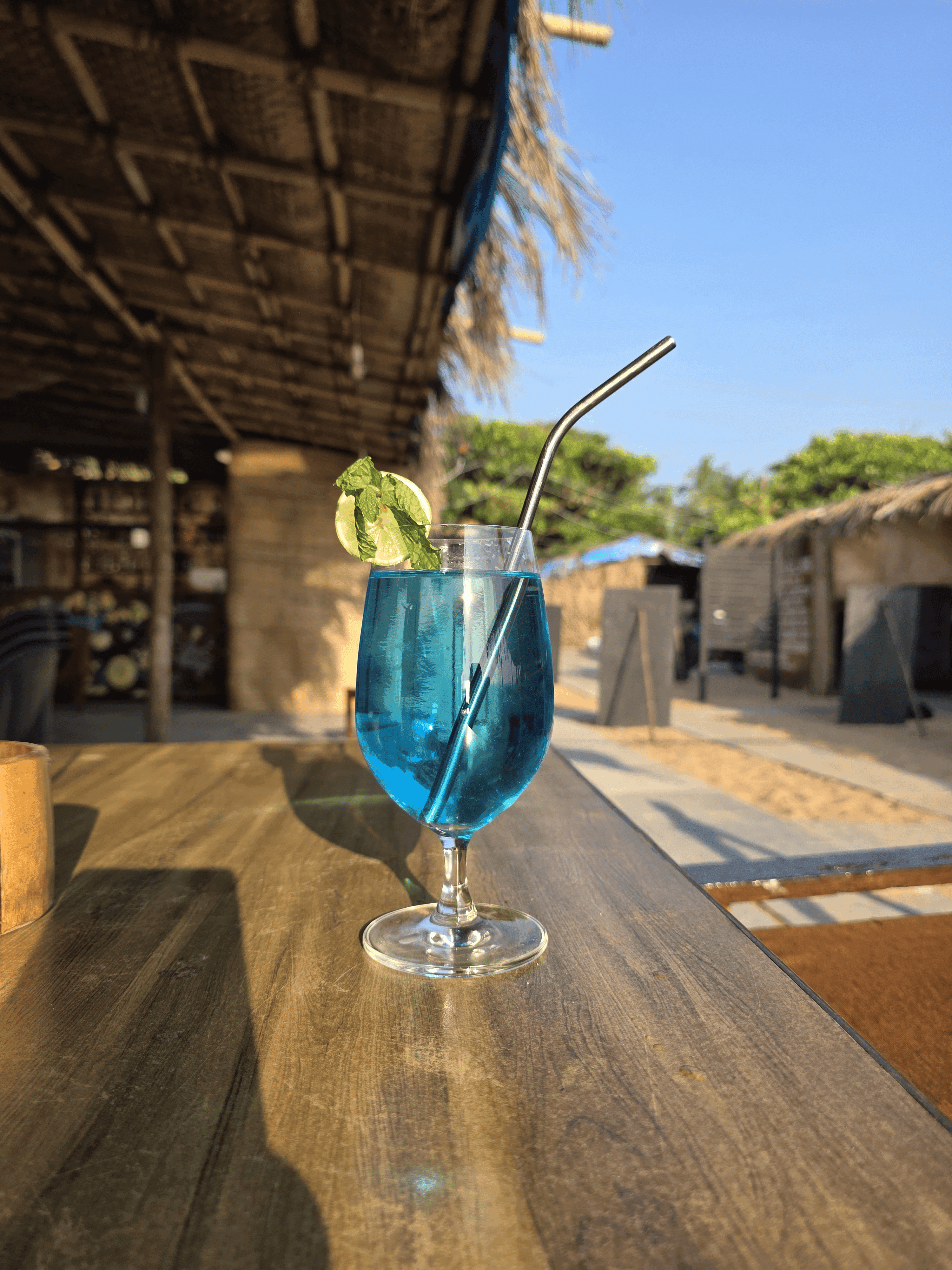 Blue lagoon cocktail with lime and mint in a stemmed glass on a wooden bar counter at golden dusk, bar and sandy path behind