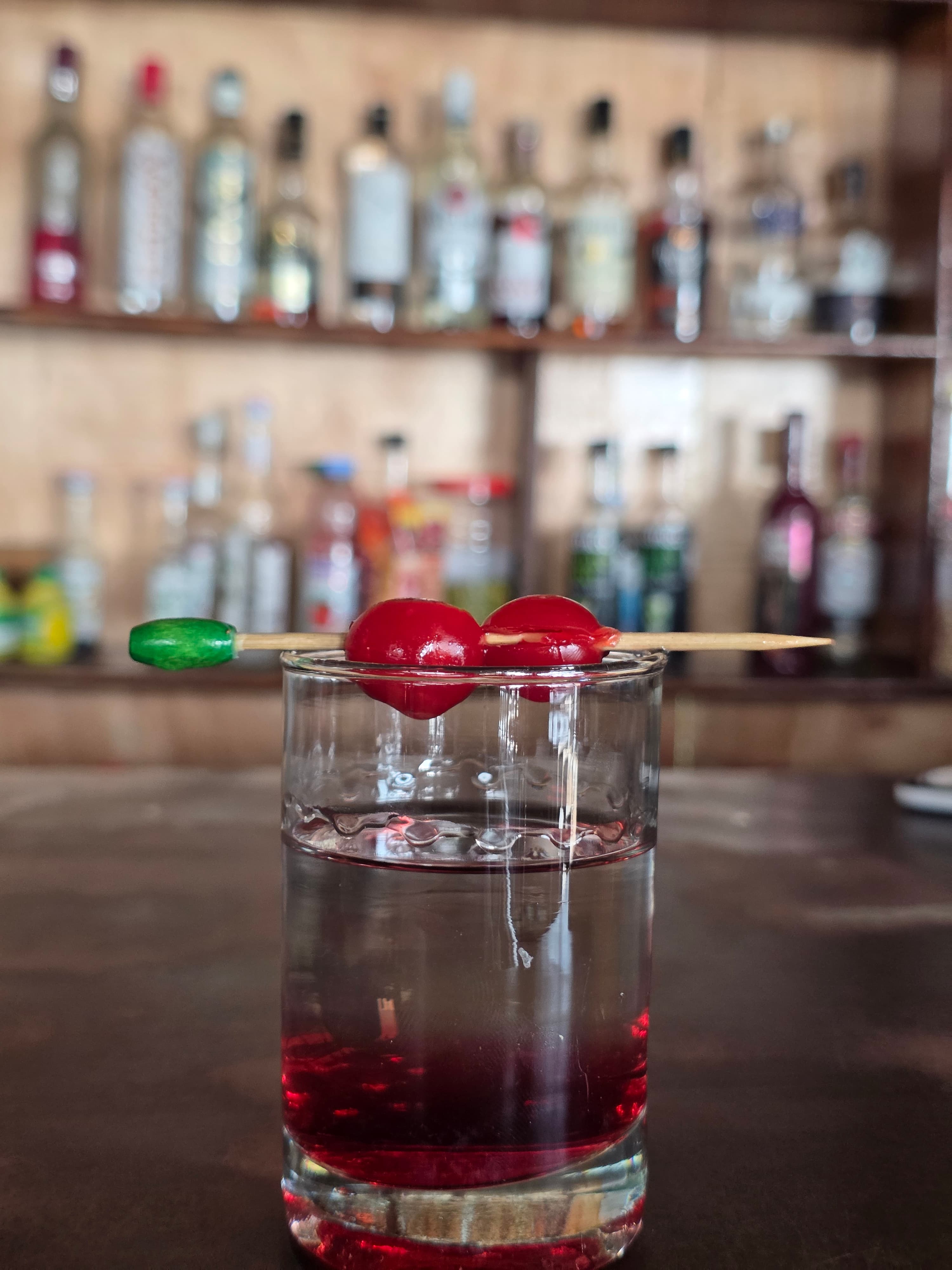 Dark crimson cocktail in a rocks glass with maraschino cherries on a cocktail stick, full spirit bar shelves behind