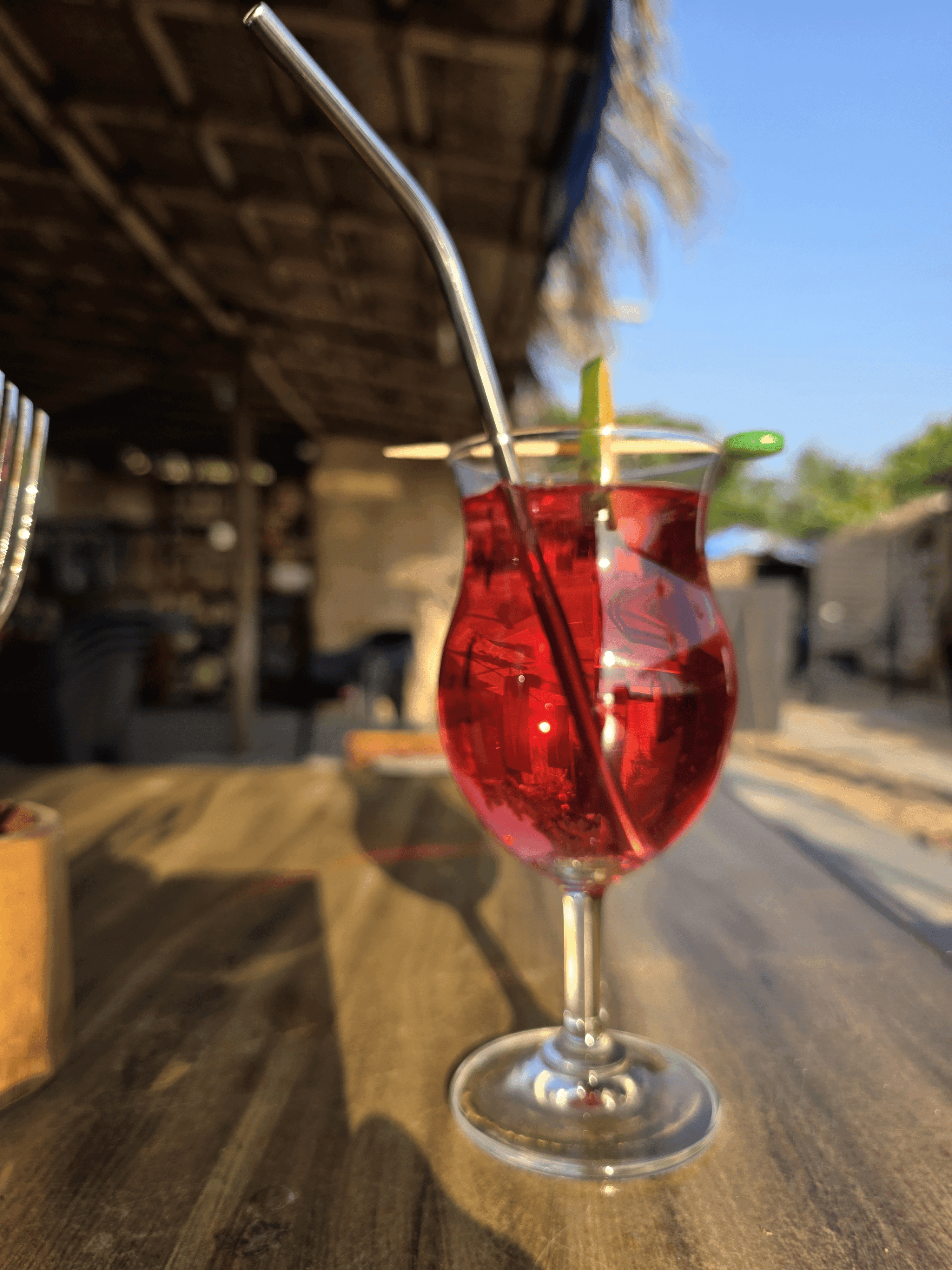 Ruby red cocktail with metal straw in an elegant stemmed glass on a wooden bar, warm golden sunset light across the scene