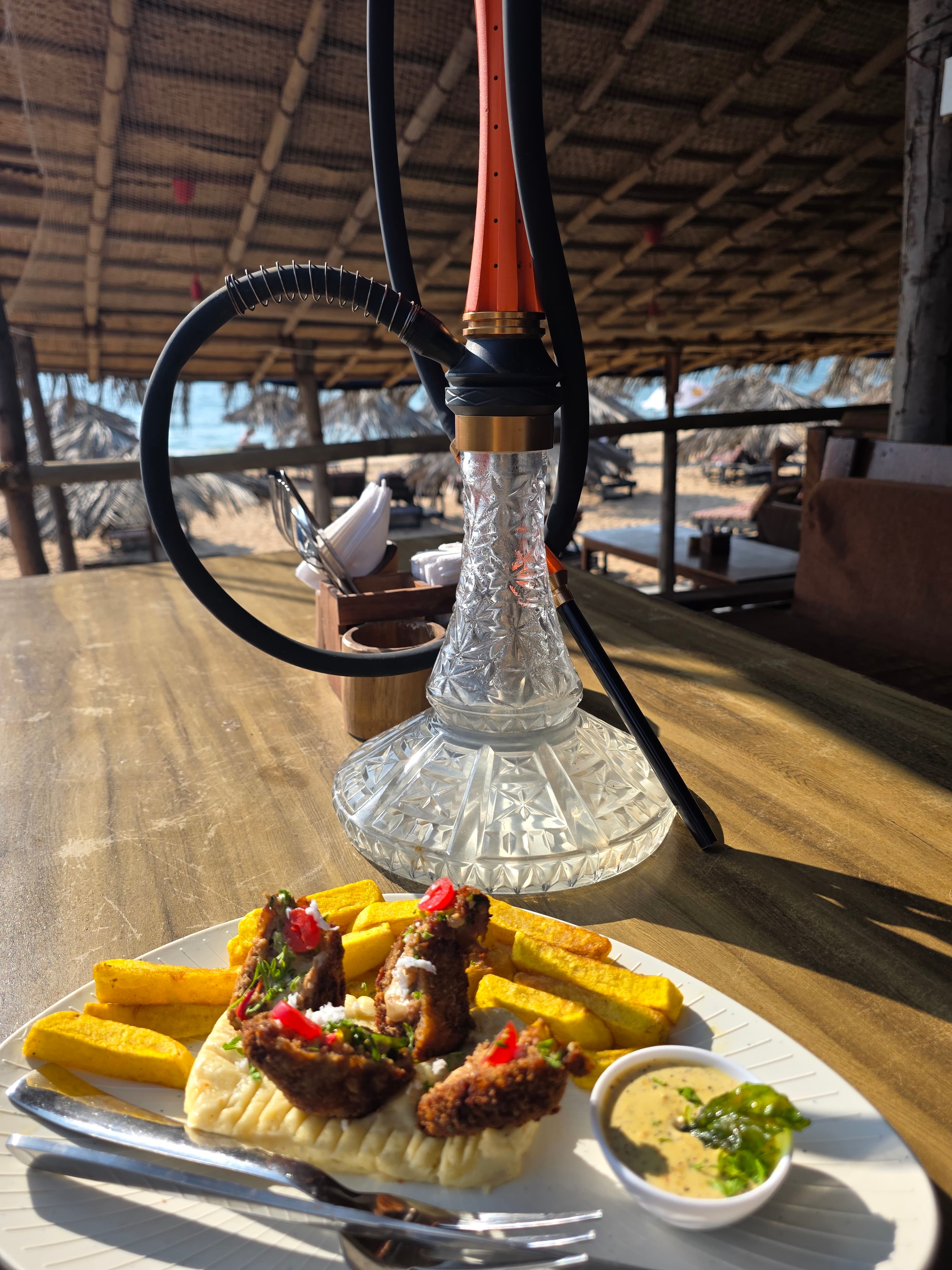 Crystal hookah shisha on a wooden table beside a plate of coastal bites, beach and thatched umbrellas in the background
