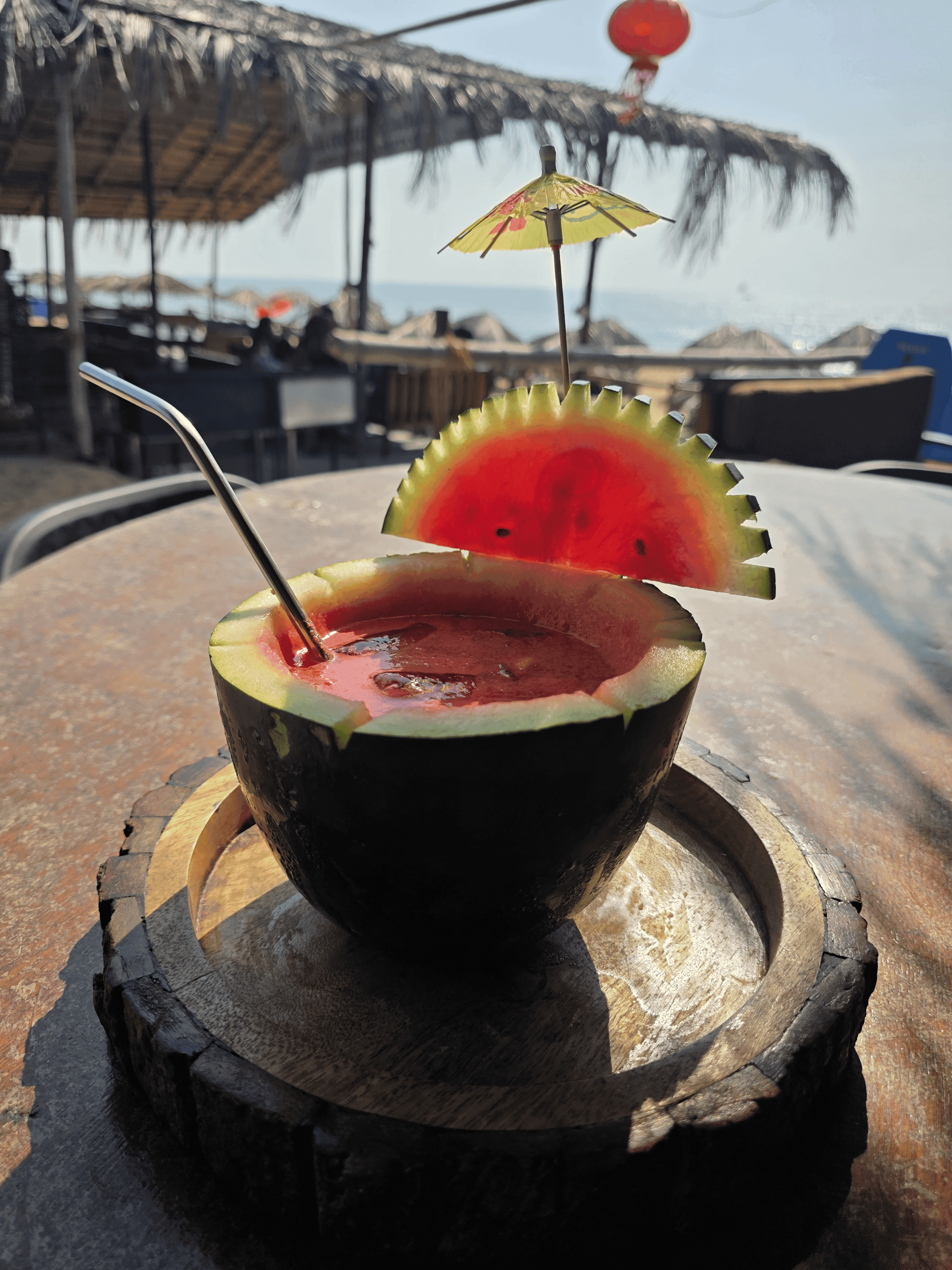Fresh watermelon cocktail served in a carved watermelon shell on a wooden platter with paper umbrella garnish, ocean behind