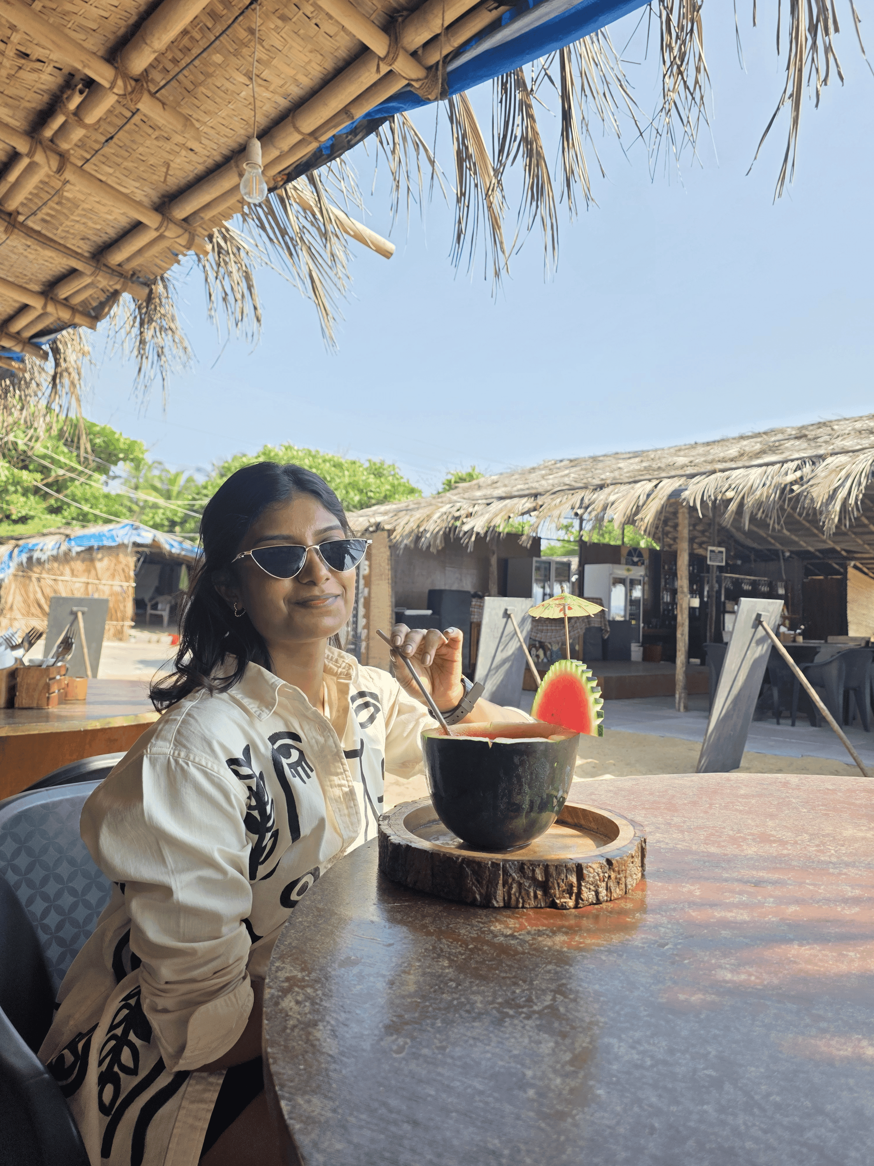 Smiling guest in sunglasses with a watermelon shell cocktail at a table under the thatched café, blue sky behind