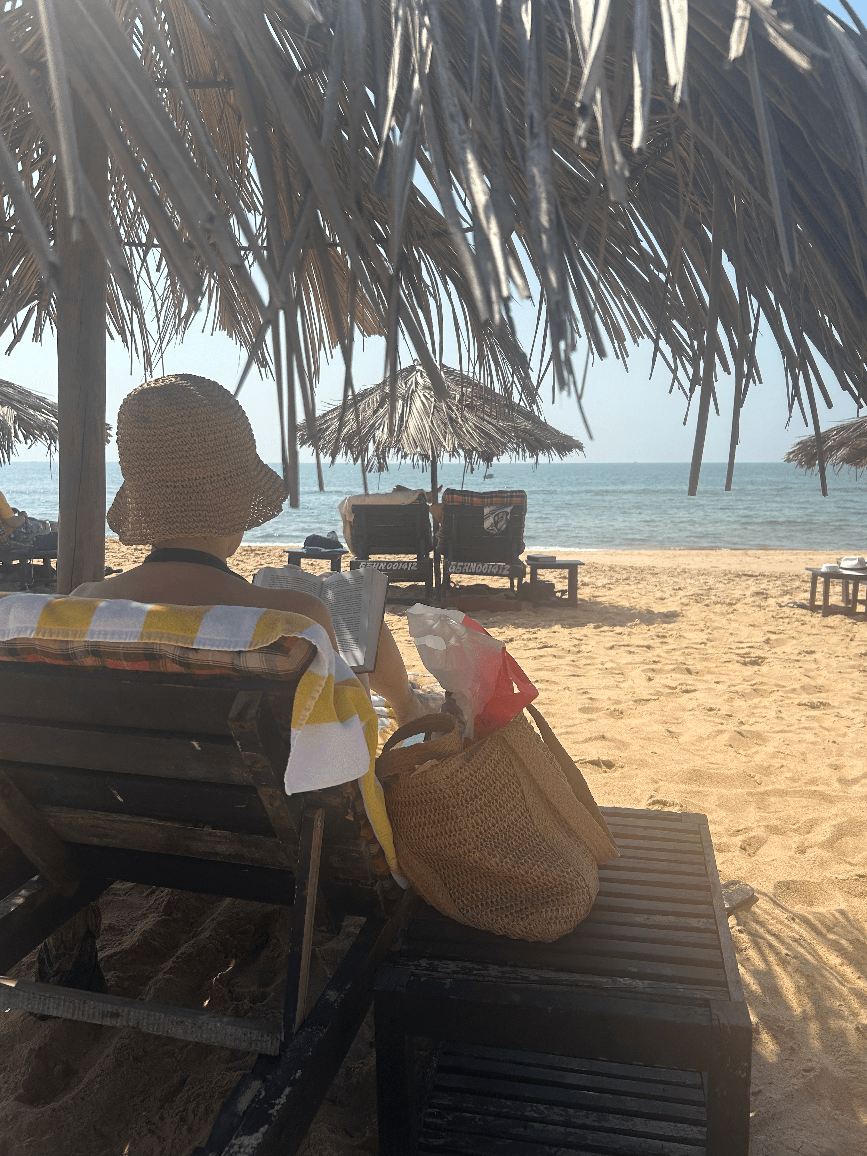 Guest in a straw hat reading on a cushioned beach lounger under a thatched parasol with the ocean in the distance