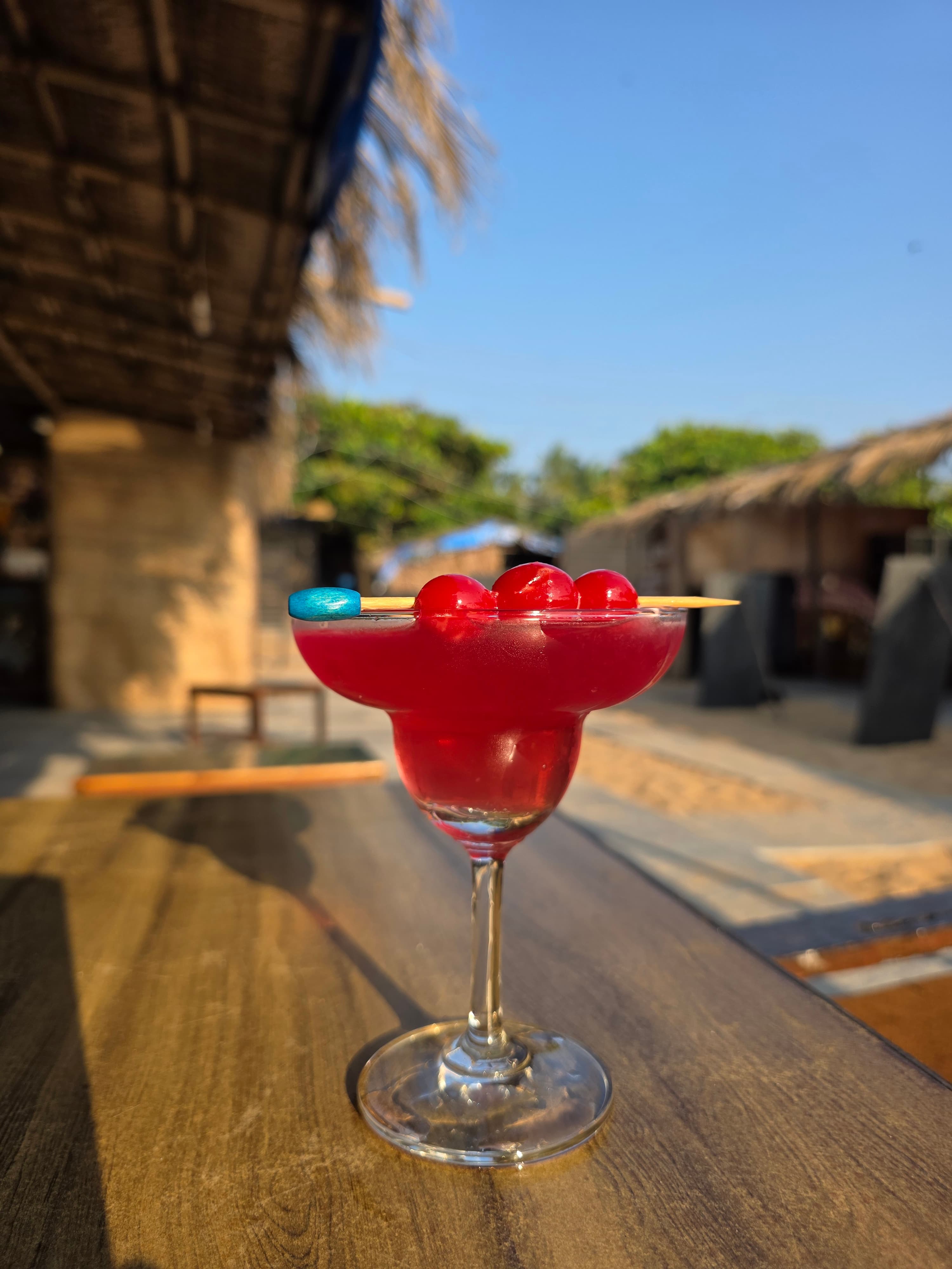 Ruby red cherry margarita cocktail in a coupe glass with maraschino cherries garnish, blue sky and thatched café behind